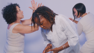 Three dancers wearing white move around each other. One hunches over, another claps, and a third leans out of the frame.