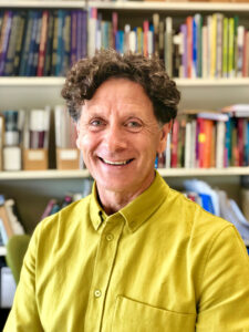 Headshot of Ralph Buck wearing a yellow shirt with bookshelves in the background.