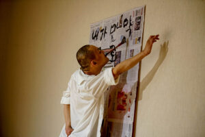 Sungkuk is painting calligraphy on a newspaper tacked on a wall. One of his hands is outstretched to the side.