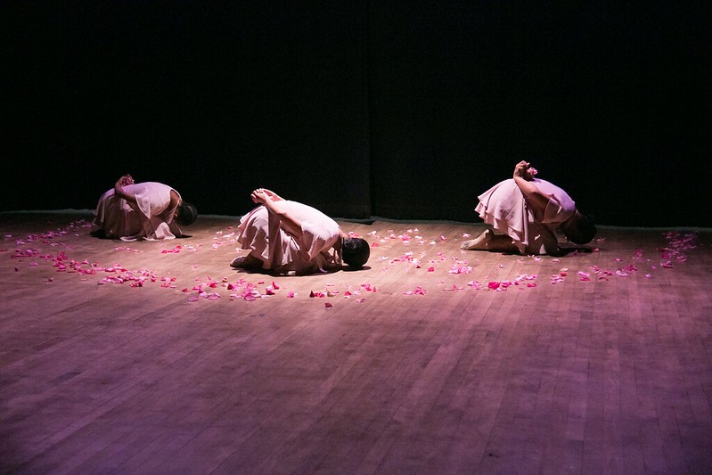Three dancers hunched over in child's pose on stage with flower petals surrounding them.