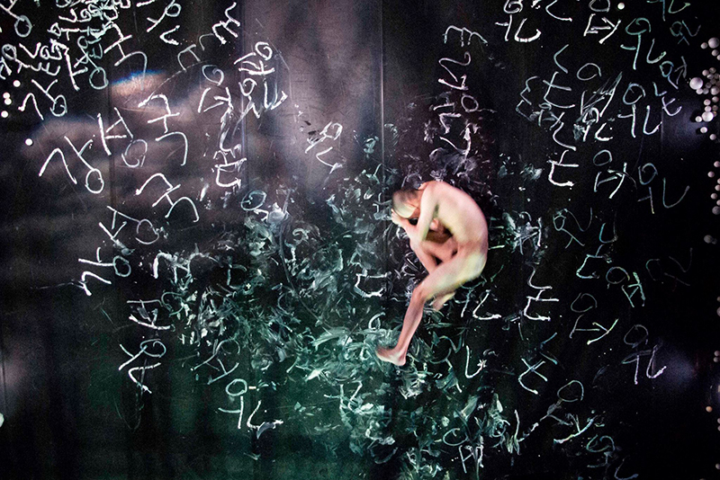 A dancer lies naked seen from above curled in a fetal-like position. Etchings like those on a chalkboard surround him.