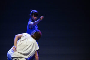 Onstage, a dancer extends her am to the side over another dancer who is lunging toward her.