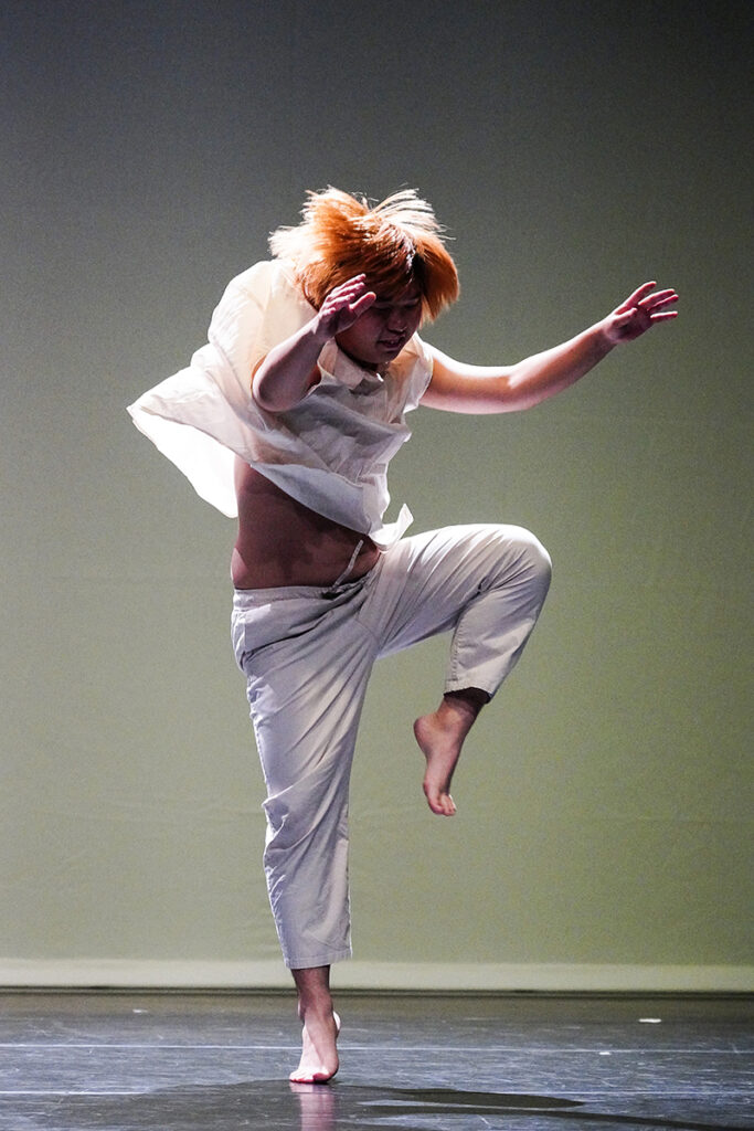 Onstage, a dancers lifts upward with his arms and one leg. He is wearing all white and his clothes move around him.