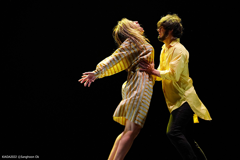 Helliot catches Laura by the ribs as she extends her arms and jumps into him. They are both wearing yellow on a black stage.