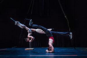 Erin and Vanessa are inverted onstage with their legs in splits. Vanessa is in the back with stilts. Erin is in the front with prosthetics.