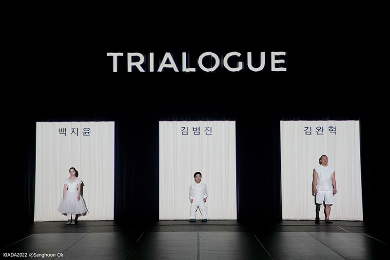 Three dancers stand in white lit rectangles onstage with the word "Trialogue" projected overhead.