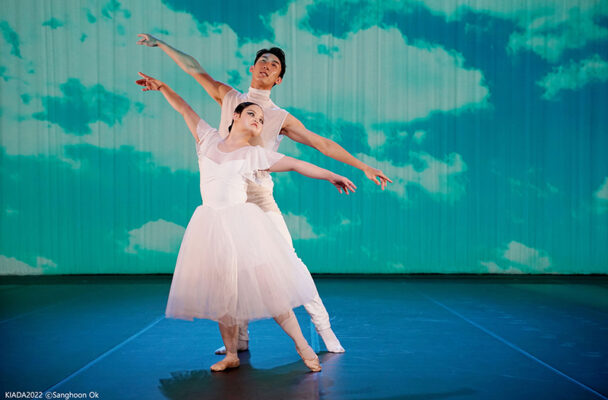 Two ballet dancers wearing white tendu to the side. The man is behind the woman. The backdrop is blue and green.