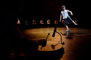 Onstage in the foreground is a microphone and drum. In the background are dancers sitting on chairs. In the middle a dancer stands with one leg lifted off the floor.
