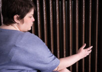 A closeup of Maggie running her hands on a radiator. She is wearing a blue shirt.