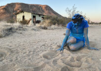 Liz sits in the sand wearing a blue outfit with her skin painted blue. A desert scene is behind her.