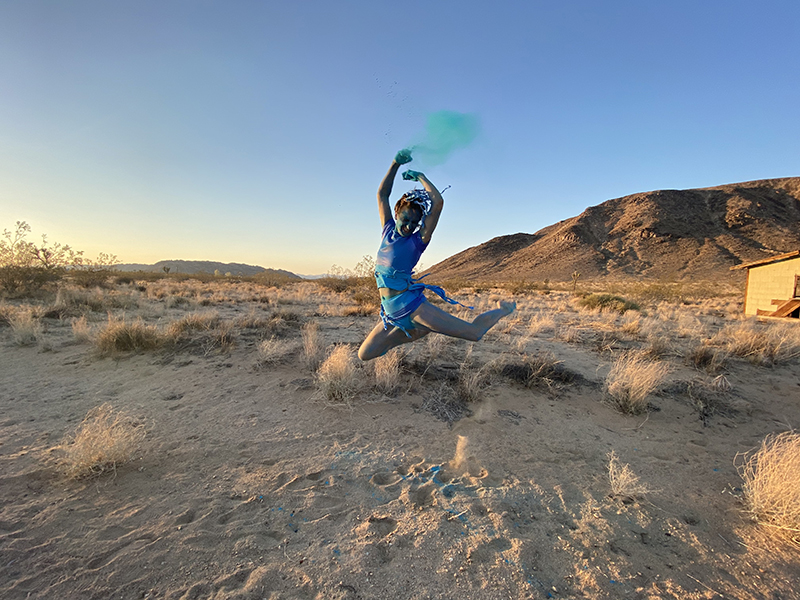 Liz leaps with her legs tucked under her. She is wearing a blue outfit and her skin is painted blue. She is in a sandy desert.