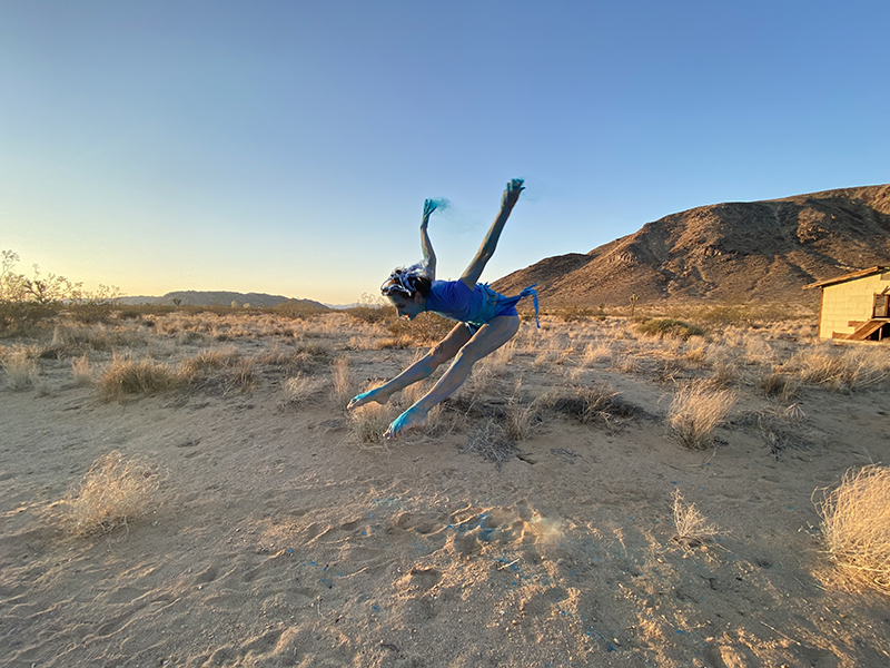 Liz leaps in a pike with her legs and arms outstretched. She is wearing a blue outfit and her skin is painted blue. She is in a sandy desert.