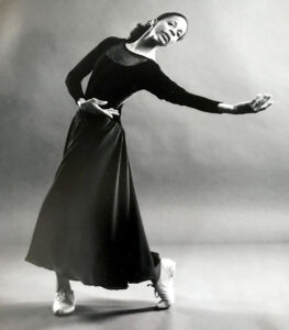 A black and white photo of Diana wearing a long black dress and leaning gently backward with an outstrethed hand.