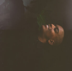 A low lit picture of breana smiling. The picture is rotating on its side. In the background you can kind of see a houseplant.