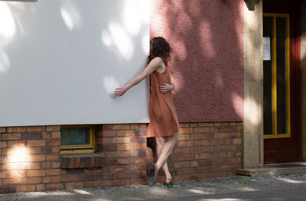 Shannon leans on a building with her hand on a white wall and her body and head turned away on a pick wall.