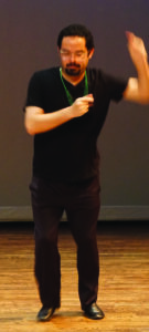 Preston stands onstage with one arm in a snap position in front of him and the other arm raised to his side.