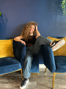 Brooke smiles while sitting on a couch with one leg on the couch arm.