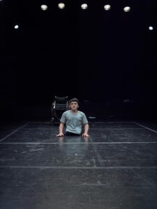 Wonyoung sits on the stage in front of his chair with both arms at his sides.