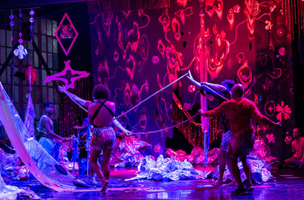 Four dancers move about on a red and silver lit stage covered with many props.