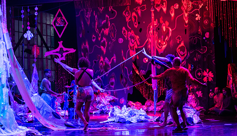Five dancers move about on a red and silver lit stage covered with many props.