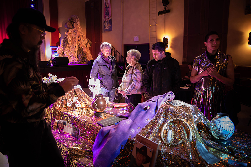 Five people mill around a table covered in gold cloth with artifacts placed on top of it.