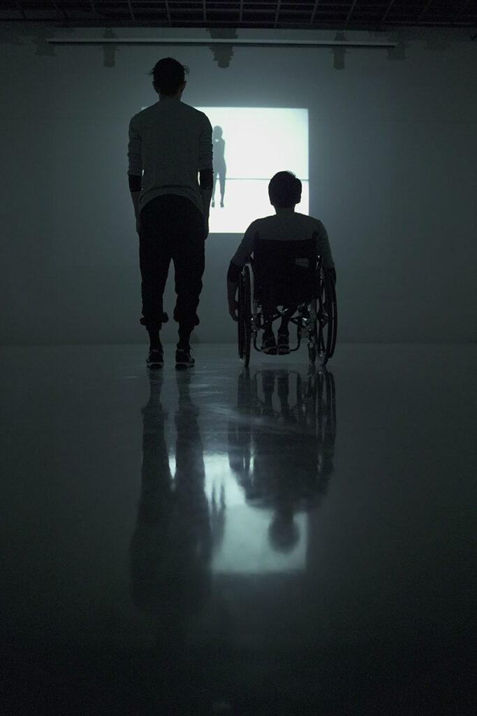 A silhouette from behind of Wonyoung in his wheelchair and a dancer standing next to him. They look at a bright screen.