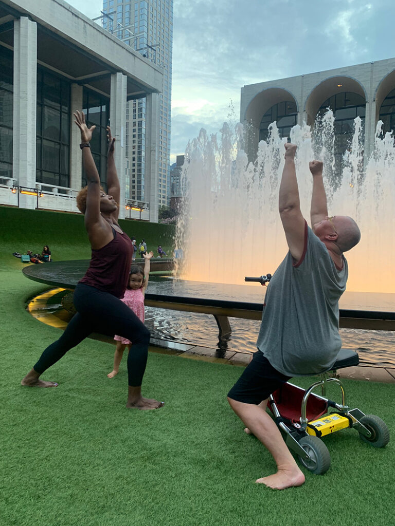 Two dancers reach upward. They are outside on a lawn with a fountain in the background.