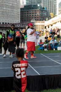 P-top emcees an event outdoors on a court with lots of people around. City buildings rise in the background.