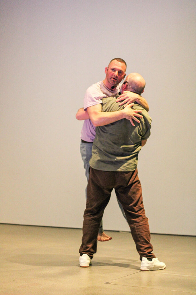Stuart and another performer are in a studio embracing each other in a hug. The other performer lifts Stuart a little off the ground.