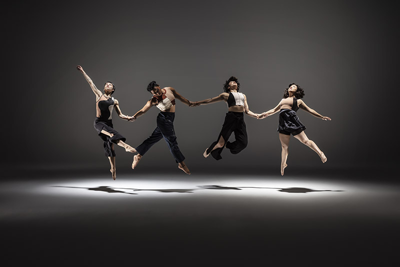 Four mixed-race Asian American dancers stretched out horizontally holding hands, jumping up in unison under a pool of light.