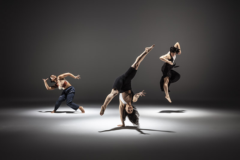 Three mixed-race Asian American dancers jump, bend, and do a handstand under pools of light.