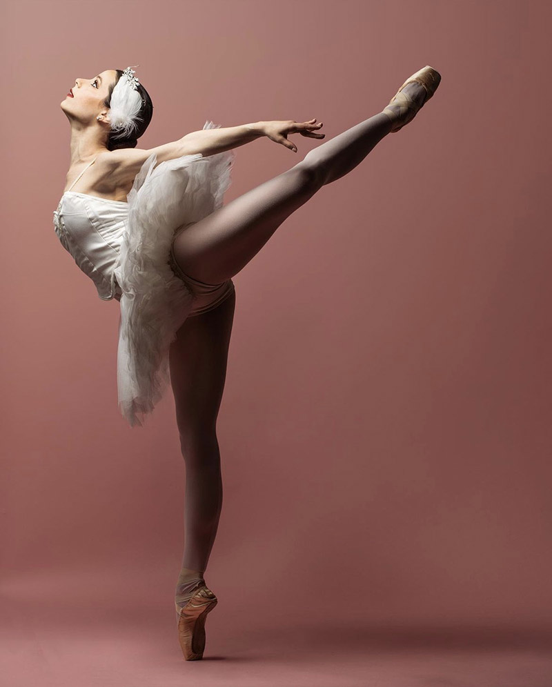 Sydney in a dramatic arabesque wearing a white swan tutu against a pink backdrop.