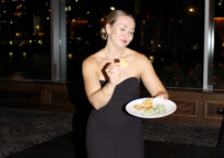 Sydney wearing a black dress and holding a plate at a party. She jokingly makes a funny pose holding a chip.