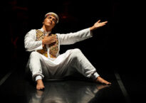 A dancer in a white tunic sits on their bottom with hands and feet bent in front of them onstage.