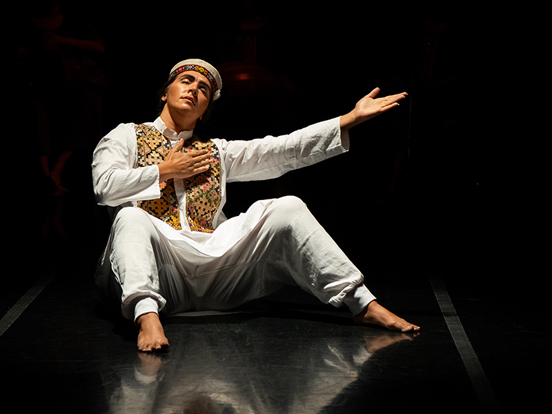 A dancer in a white tunic sits on their bottom with hands and feet bent in front of them onstage.