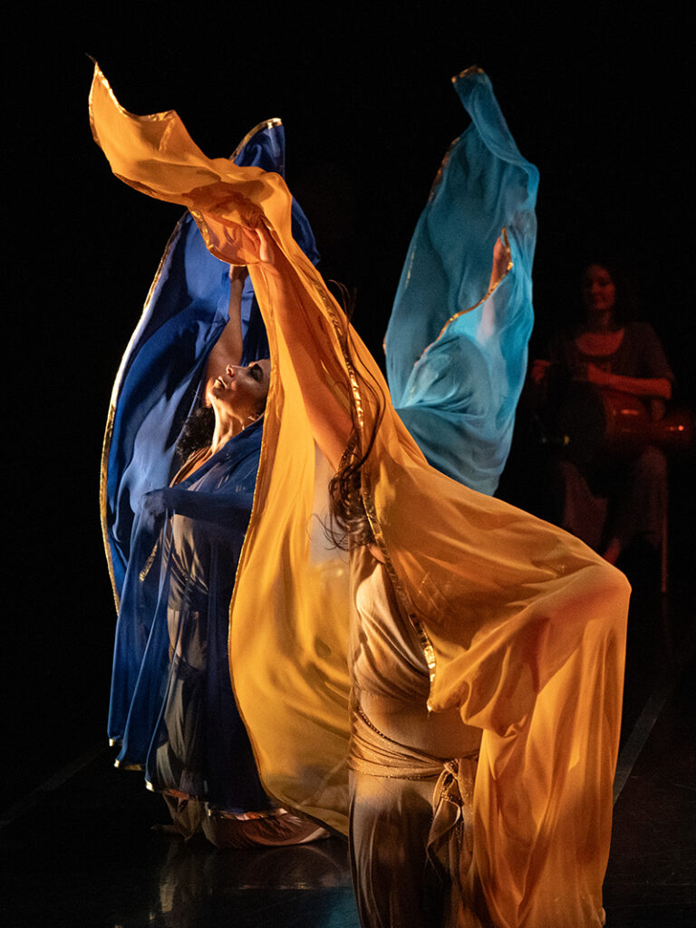 Three dancers with bright scarves reach upward and project the scarves above them.