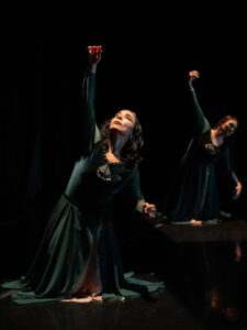 Two women kneel onstage in dark long dresses with one arm extended upward.
