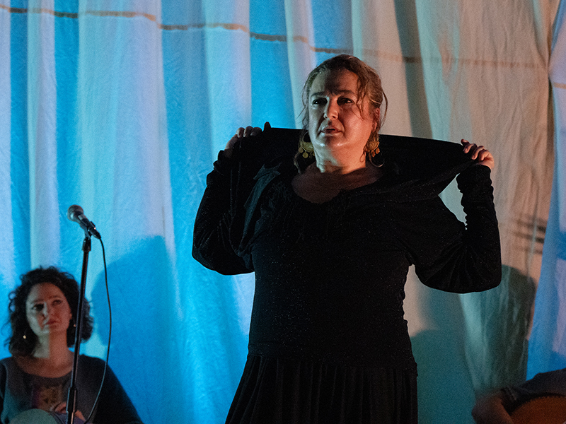A woman in black with a curtain and microphone behind her reaches behind her as if to take off a jacket.
