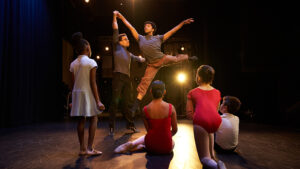 Melendez aids a student in a leap as other students look on. They are on stage.