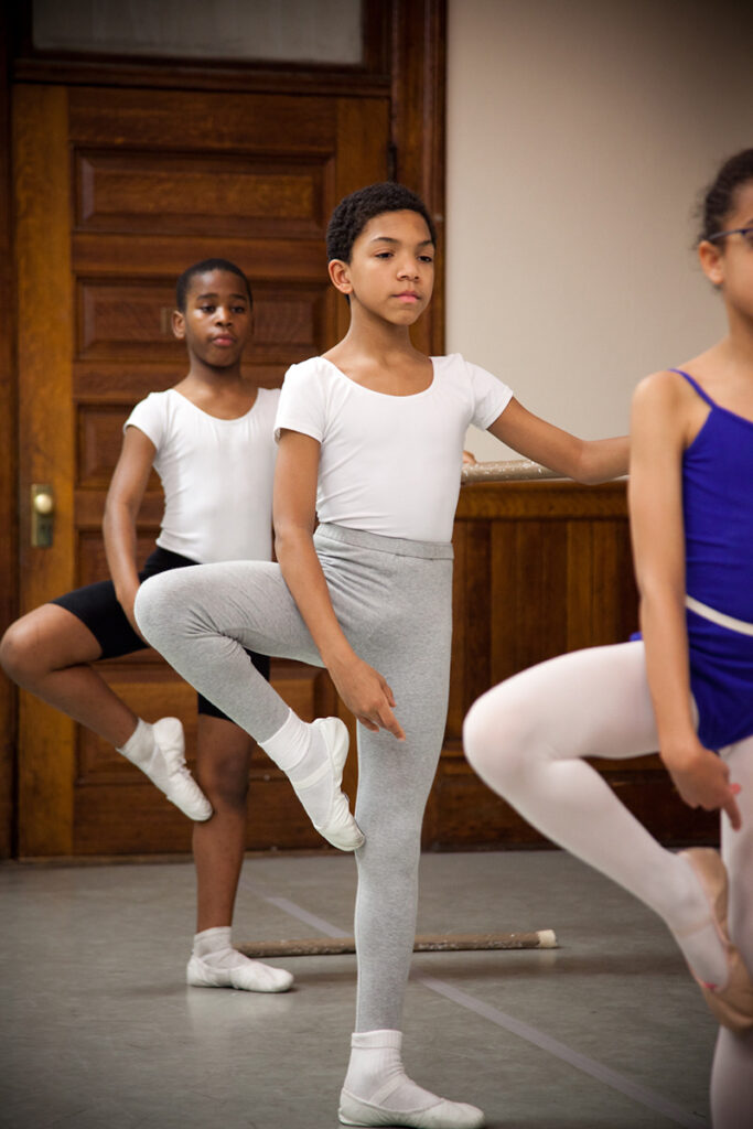 Children stand along a barre in a ballet studio practicing passe.