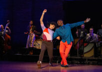 Two dancers link arms and lean back. Behind them in a live onstage band.