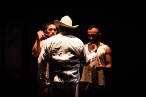 A person in a big hat onstage with his back to the audience faces to other performers.