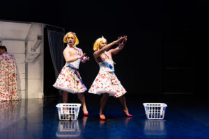 Two performers wear blond wigs and skirts with laundry baskets nearby.