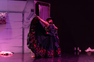 Rogelio on stage with arms reaching out to his sides, wearing a black frilly dress.