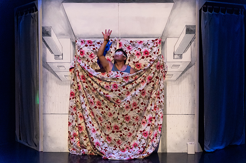 Rogelio onstage with flowery sheets draped around him in the shape of a bed.