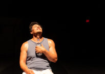 Rogelio sits on stage in a tank top and looks upward with a water bottle to his chest.