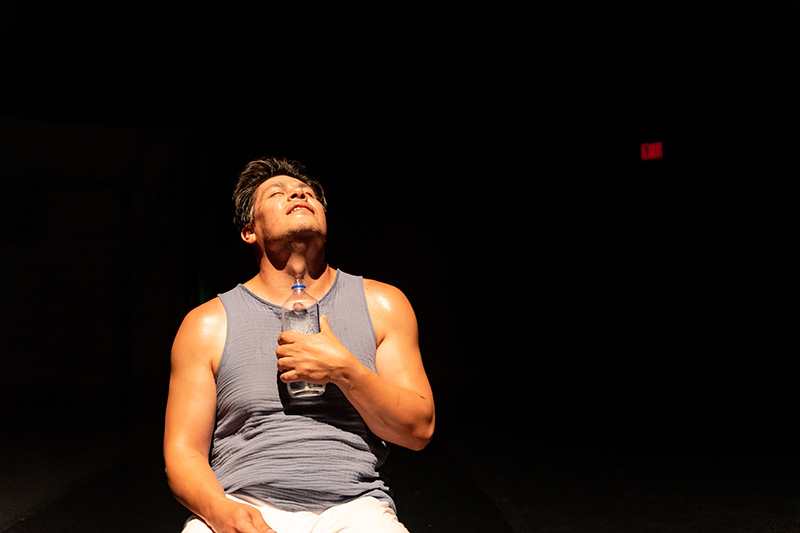 Rogelio sits on stage in a tank top and looks upward with a water bottle to his chest.