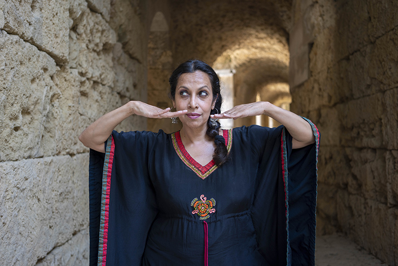 Shebana stands in a stone tunnel. She is wearing a black tunic. Her hands are level above her chin and she looks to the side.