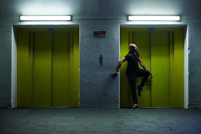 Darrell dances in front of one of two elevators with green doors in a parking garage.