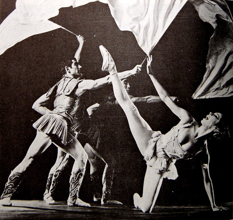 An old photo of a ballet dancer on her knee with one leg in the air. Other dancers lunge behind her.